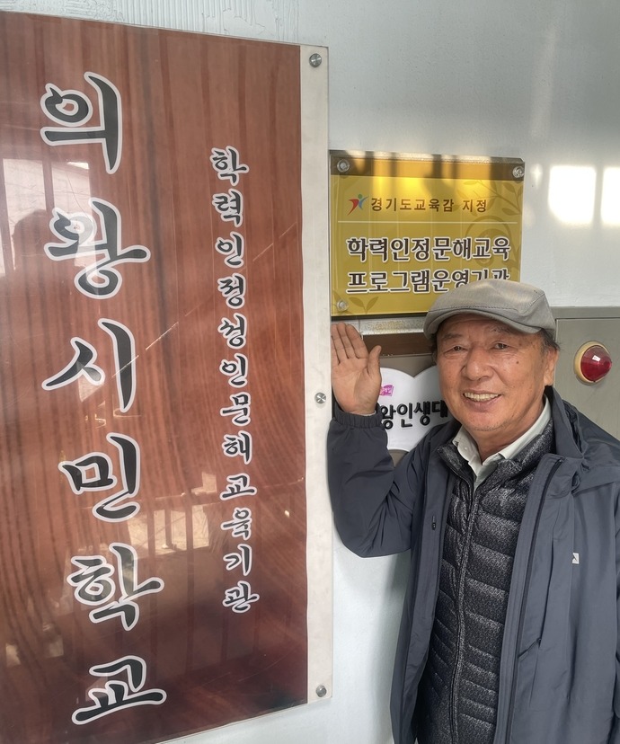 이웅기 의왕시민학교 교장이 ‘학력인정 문해교육 프로그램 운영기관’ 현판 앞에서 밝게 미소 짓고 있다. 손용현기자